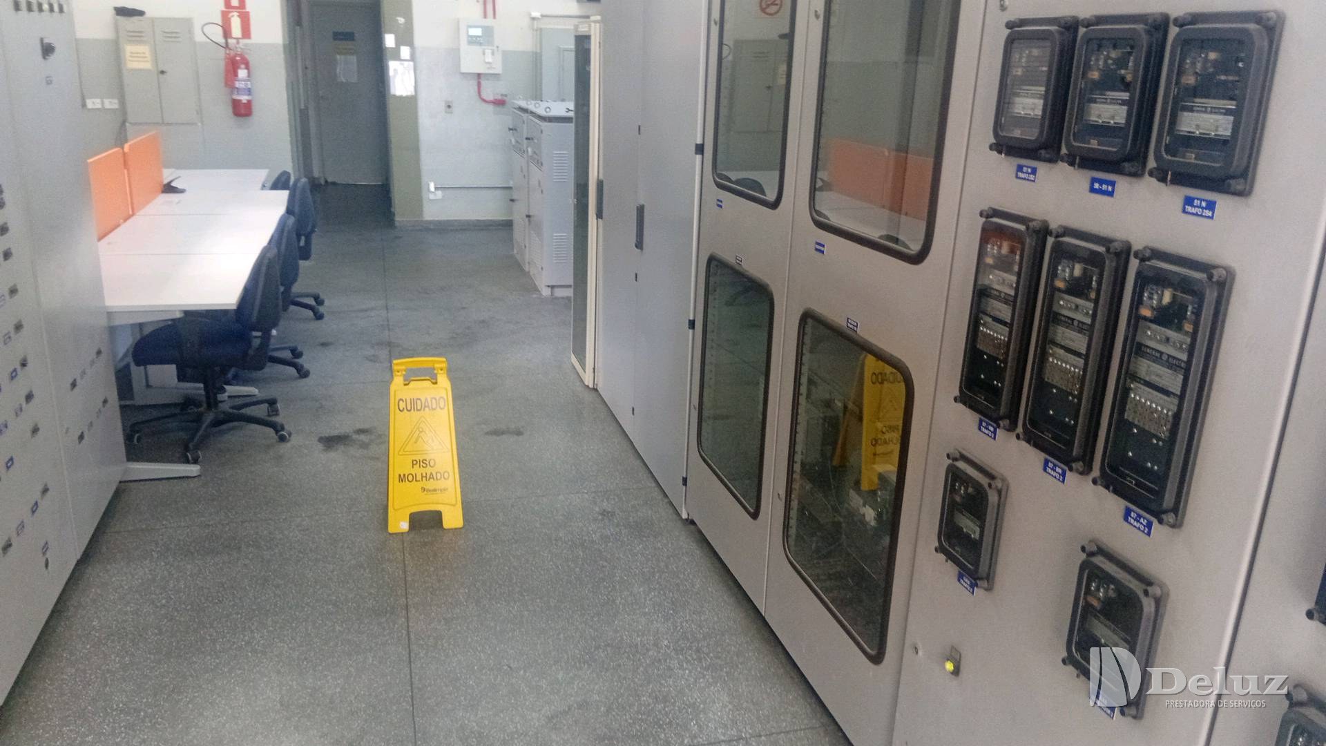 Sala de comando com painéis elétricos e sinalização de piso molhado durante a limpeza