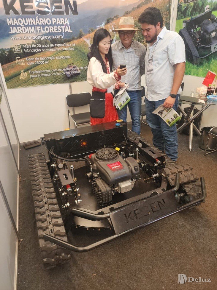 Máquina de roçada sobre esteiras exposta em estande de feira, com três pessoas ao redor