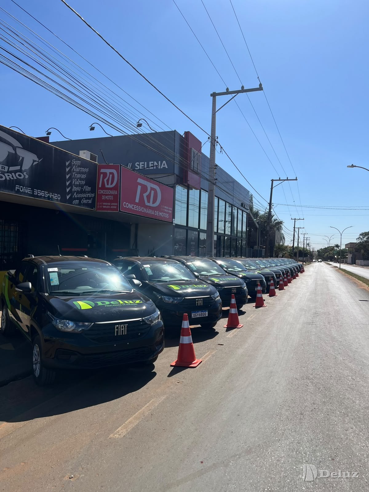 Fila de Fiat Strada pretas alinhadas na rua, em frente à concessionária, sinalizadas com cones